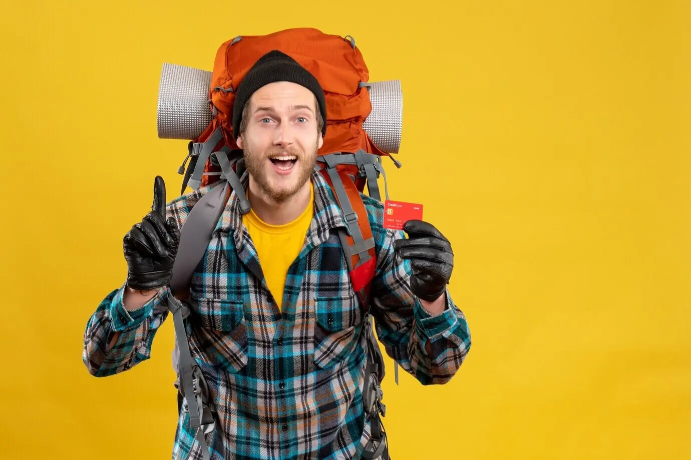 Front view of a cheerful camper with a backpacker, holding a credit card and pointing at the ceiling.