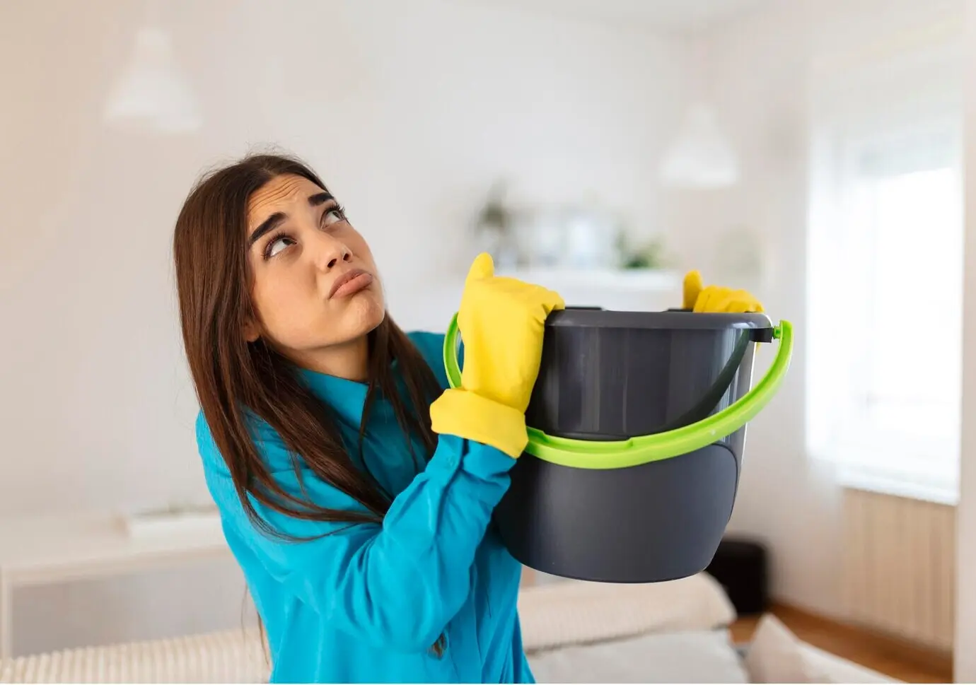 A shocked, worried woman looks up at the ceiling in the living room at home, holding a bucket to collect water as droplets leak from the ceiling.