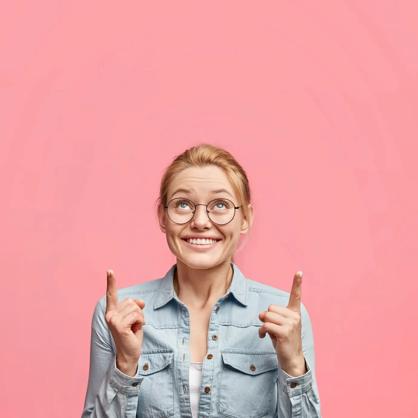Vertical shot of a delighted, pretty young blonde woman with a pleasant appearance, pointing upward with a happy expression, advertising something on the ceiling.